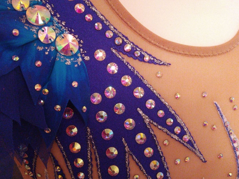 May include: A close-up of a purple leotard with a blue and teal feather design. The leotard is decorated with iridescent rhinestones.