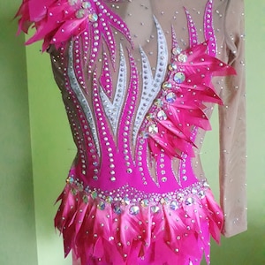 May include: A pink leotard with a white and silver design, adorned with rhinestones and feathers. The leotard has long sleeves and a ruffled skirt.
