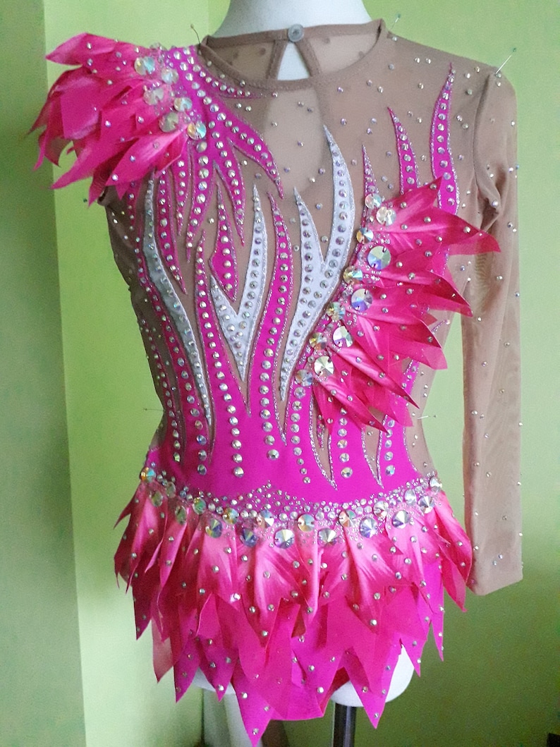 May include: A pink leotard with a white and silver design, adorned with rhinestones and feathers. The leotard has long sleeves and a ruffled skirt.