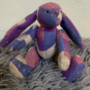 May include: A patchwork bunny plush toy in shades of purple, pink, and cream. The bunny has long ears, a small brown nose, and is sitting on a fluffy gray surface. The toy is made of fabric pieces sewn together.