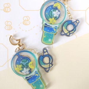 Könnte beinhalten: Zwei Schlüsselanhänger aus Acryl mit einem himmlischen Thema. Jeder Schlüsselanhänger hat ein blaues und grünes Design mit Sternen, Monden und einem Planeten. Einer hat die Form eines Bubble Tea, der andere ein Planeten- und Sicherheitsnadel-Design. Beide haben goldfarbene Hardware.