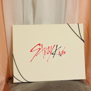 Könnte beinhalten: Cremefarbene rechteckige Box mit dem Schriftzug "Stray Kids" in roter und schwarzer Schrift. Schwarze Ketten akzentuieren die Ecken der Box. Die Box liegt auf einer grauen Oberfläche, mit einem rosa Vorhang im Hintergrund.