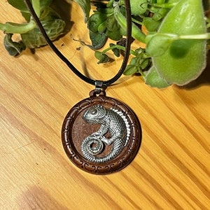 May include: A round pendant necklace featuring a silver chameleon design on a brown background, set within a decorative brown frame. The pendant hangs from a black cord. The background includes greenery and a wooden surface.