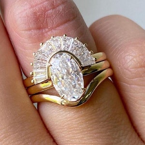 Puede incluir: Un anillo de oro engastado con un gran diamante de forma ovalada rodeado de diamantes baguette m&aacute;s peque&ntilde;os. El anillo se lleva en un dedo con una banda de oro a juego.