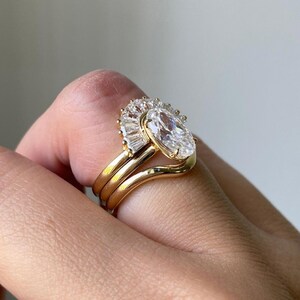 Puede incluir: Un anillo de oro con un gran diamante de forma ovalada rodeado de diamantes baguette m&aacute;s peque&ntilde;os. El anillo est&aacute; montado en una banda curva y se lleva en un dedo.