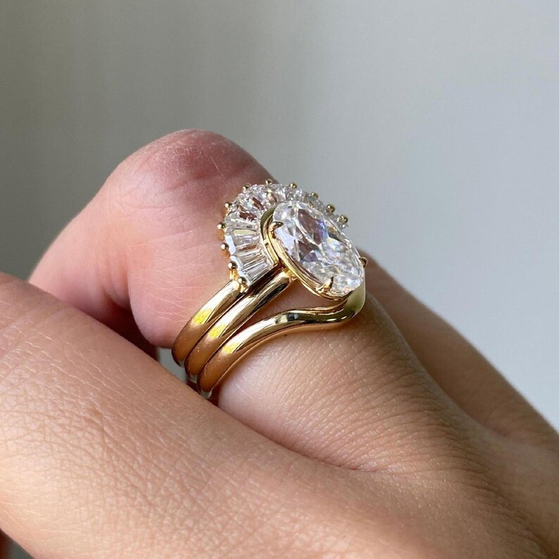 Puede incluir: Un anillo de oro con un gran diamante de forma ovalada rodeado de diamantes baguette m&aacute;s peque&ntilde;os. El anillo est&aacute; montado en una banda curva y se lleva en un dedo.