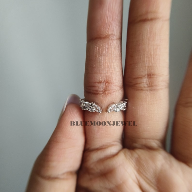 May include: A silver ring with a leaf design and clear stones. The ring is open in the back and is adjustable to fit different finger sizes.  The ring is on a finger.