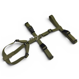 May include: An olive green nylon pet harness featuring adjustable straps, black plastic side-release buckles, and metal D-rings for leash attachment. The harness includes a small black fabric tag with white text attached to the central strap.