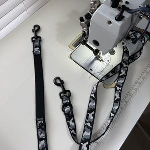 Puede incluir: Dos correas para perro con un patrón de camuflaje en negro, gris y blanco. Cada correa incluye un mosquetón giratorio de metal negro y una anilla en D negra. Una correa es parcialmente negra sólida y una regla muestra medidas en centímetros.