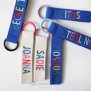 Puede incluir: Cinco llaveros personalizados coloridos con diferentes nombres bordados. Los llaveros están hechos de tela azul y blanca con anillos negros, rosas y plateados. El texto en los llaveros dice "Beckett", "Titus", "Sterling", "Joanna", "Sadie" y "Joshua".