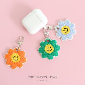 May include: Three fuzzy flower-shaped keychains with smiling faces. The keychains are blue, green, and orange. The text "THE LEMONG STORE HANDMADE" is at the bottom of the image.