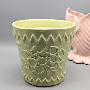 May include: A light green ceramic planter with a zigzag pattern around the rim and a floral design. A pink ceramic decorative piece is partially visible in the background. The planter is cylindrical with a wide opening.