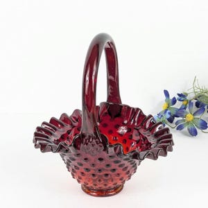 May include: A ruby red glass basket with a handle. The basket has a ruffled rim and a hobnail pattern. The handle arches gracefully. The basket is likely vintage and decorative.