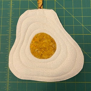 May include: A handmade, quilted egg-shaped potholder. The potholder features a white egg white with a yellow yolk. The egg white has a subtle floral pattern and is stitched with concentric circles. A small loop is attached at the top.