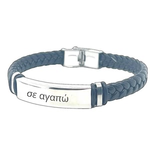 Puede incluir: Pulsera de cuero negro con una placa plateada grabada con la frase griega "σε αγαπώ" (Te amo).