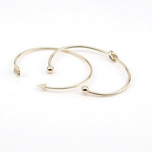 May include: Two gold-toned bangle bracelets. One bracelet features a small arrow charm and the other has a knot design.