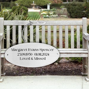 Może przedstawiać: Drewniana ławka parkowa z tablicą pamiątkową. Na tablicy napis: "Margaret Evans Spencer 23.09.1950-01.01.2024 Kochana i wspominana". Ławka jest jasnoszara i znajduje się w ogrodzie z fontanną.