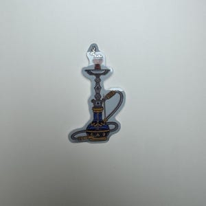 May include: A sticker of a hookah with a blue base, gold accents, and a silver stem. The top of the hookah has a red bowl with smoke rising. The hose is silver and curved. The sticker is on a white background.