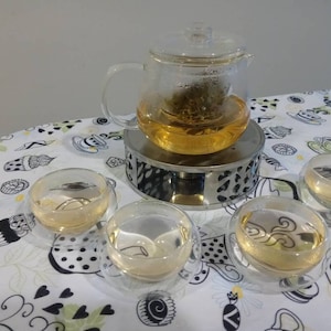 May include: A glass teapot with a built-in infuser sits on a warming base, surrounded by four clear glass teacups filled with light-colored tea. The teacups are on a white tablecloth with a black and white pattern.