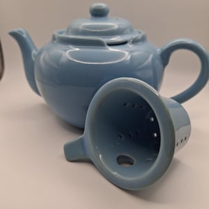 May include: A light blue ceramic teapot with a matching infuser. The teapot has a rounded body, a curved spout, and a loop handle. The infuser is cone-shaped with small holes for tea leaves. The lid has a small knob.