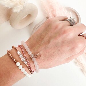 Peut inclure: Un ensemble de cinq bracelets avec des perles et des breloques en or rose. Les bracelets comprennent un bracelet à perles blanches avec un pendentif en forme de cœur, un bracelet à perles roses, un bracelet à perles transparentes avec le mot "MAMA" écrit, et deux bracelets à perles en or rose.
