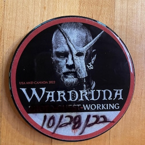 May include: Round black button with a red border featuring a grayscale image of a man wearing a mask. The text "Wardruna" is displayed in white, along with "USA AND CANADA 2022" and "PHOTO GUEST-WORKING". The date "10/28/22" is handwritten.