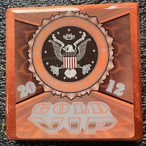 May include: A red, white, and blue American flag with an eagle and the words "American Capital" and "2012" on a brown background with the words "Gold VIP" in silver.