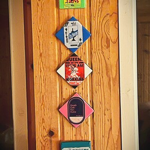 May include: Five square-shaped, colorful, framed prints with band names and album titles. The prints are arranged vertically on a wooden wall. The top print is green with the band name "MANESKIN" and the album title "LOUD KOY TANR". The second print is blue with the band name "THE" and the album title "FO FIGHTERS". The third print is white with the band name "QUEEN OF THE STONE AGE" and the album title "WORKING". The fourth print is pink with the band name "Death Cab For Cutie" and the album title "WORKING". The bottom print is teal with the band name "weezer" and the album title "SEA 4/9: WORKING".