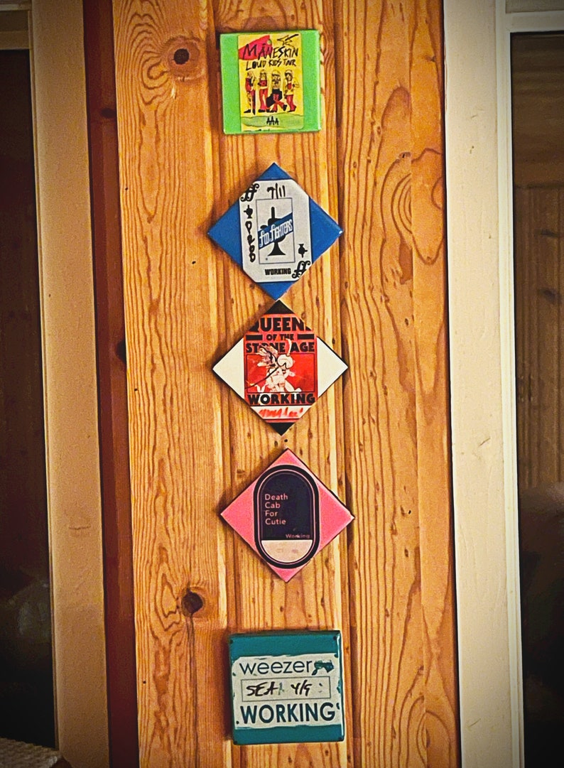 May include: Five square-shaped, colorful, framed prints with band names and album titles. The prints are arranged vertically on a wooden wall. The top print is green with the band name "MANESKIN" and the album title "LOUD KOY TANR". The second print is blue with the band name "THE" and the album title "FO FIGHTERS". The third print is white with the band name "QUEEN OF THE STONE AGE" and the album title "WORKING". The fourth print is pink with the band name "Death Cab For Cutie" and the album title "WORKING". The bottom print is teal with the band name "weezer" and the album title "SEA 4/9: WORKING".