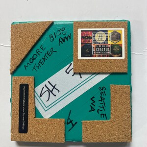 May include: A teal blue square coaster with cork backing. The coaster has black marker writing on it that says "SHE" and "Seattle WA". The coaster also has a sticker on it that says "Cuz life like rock n roll is messy and some days we all need a coaster. HeyImWithTheBand 19-29 Sea".