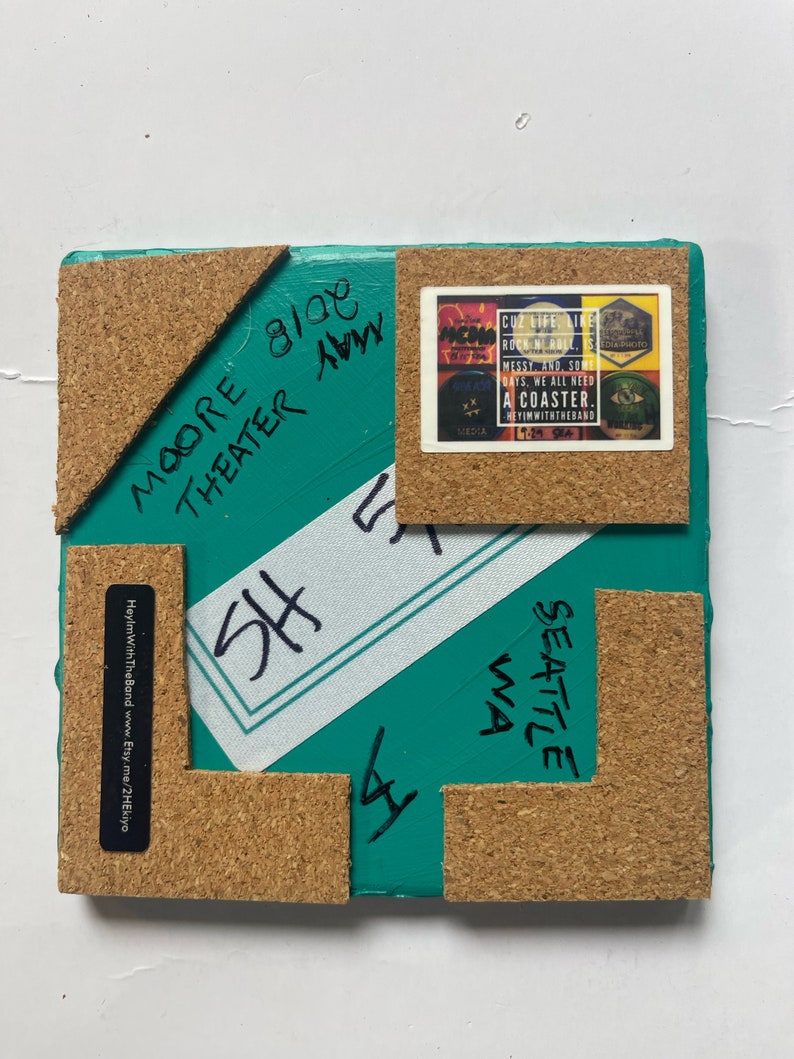 May include: A teal blue square coaster with cork backing. The coaster has black marker writing on it that says "SHE" and "Seattle WA". The coaster also has a sticker on it that says "Cuz life like rock n roll is messy and some days we all need a coaster. HeyImWithTheBand 19-29 Sea".