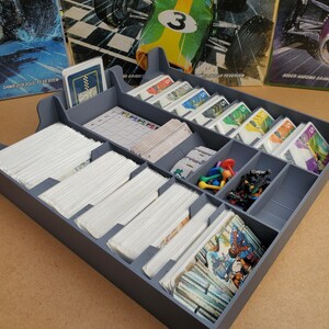 May include: A gray, compartmentalized game organizer filled with stacks of white cards, game pieces, and colorful cards. The organizer has multiple sections for different game components. The background includes game boxes with text.