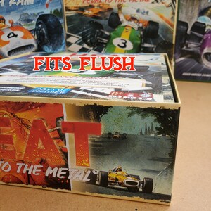 May include: A board game box with the text "FITS FLUSH" in red. The box features the title "HEAT: Pedal to the Metal" with images of race cars. Other game boxes are visible in the background, with images of race cars.