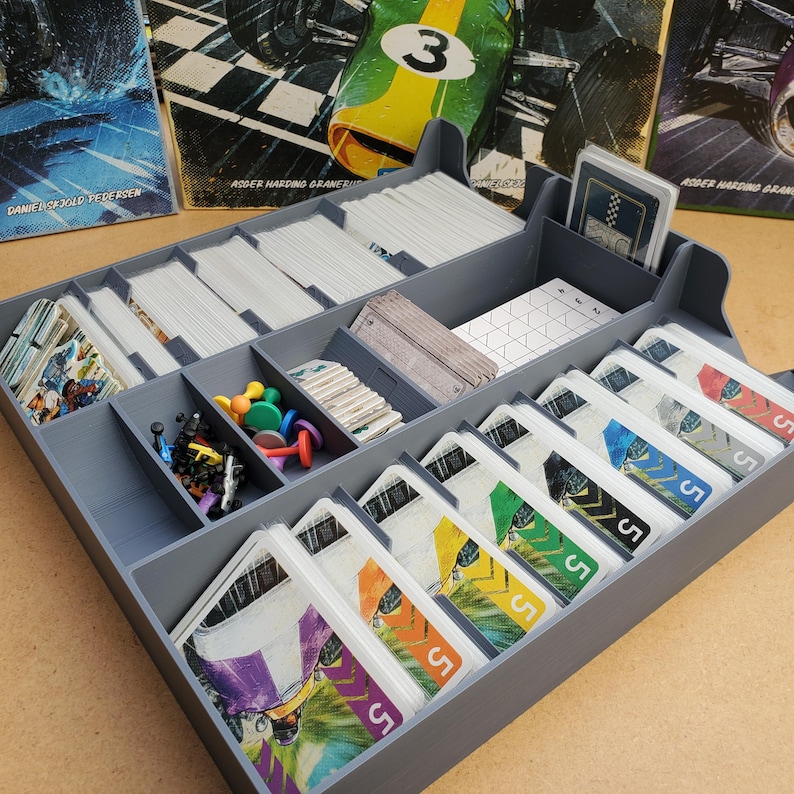 May include: A gray plastic game organizer with multiple compartments filled with game cards, tokens, and other game components. The cards have colorful racing car graphics and the number "5". The organizer is designed to hold board game pieces.
