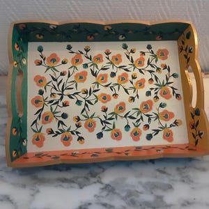 Puede incluir: Una bandeja de servir de madera con un diseño floral pintado a mano. La bandeja tiene un borde verde, un centro blanco con flores naranjas y amarillas y un borde inferior de color melocotón.