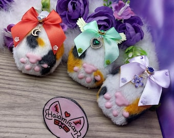 May include: Three plush cat paw keychains with different colored bows. The first keychain has a red bow with a crystal charm, the second keychain has a green bow with a green crystal charm, and the third keychain has a purple bow with a blue crystal charm.