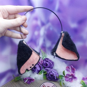 May include: Black furry cat ears headband with a pink interior. The headband is made of black wire and has a pink heart on the side. The headband is on a white fluffy surface with purple flowers.