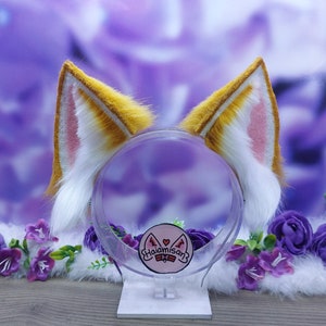 May include: A pair of white and yellow cat ears headband with pink inner ears. The headband is on a clear plastic stand with a pink and white logo that says "Haiamisanz".