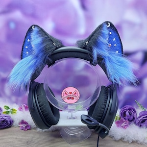 Könnte beinhalten: Schwarzes Gaming-Headset mit Katzenohren. Die Ohren sind schwarz mit blauem Kunstfell und Stern- und Mondakzenten. Das Headset wird auf einem transparenten Ständer mit Logo präsentiert.