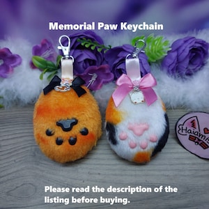 Peut inclure: Deux porte-clés en forme de patte de chat moelleux, l'un orange et l'autre calicot, avec des accents noirs et roses. Les porte-clés sont attachés à des anneaux de clés argentés. Le texte "Memorial Paw Keychain" est en haut de l'image. Le texte "Haiamisan?" est sur un autocollant rose avec un cœur et un motif de papillon.