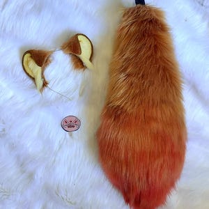Puede incluir: Un conjunto de cosplay con una cola esponjosa, naranja y roja degradada, y una diadema con orejas marrones y amarillas a juego. También hay una pequeña pegatina redonda con texto y una cara de gato.