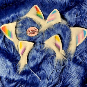 May include: White cat ear headbands with rainbow and pastel accents. Each ear is trimmed with fluffy white fur and attached to a silver headband. A pink patch with Japanese text is also visible.