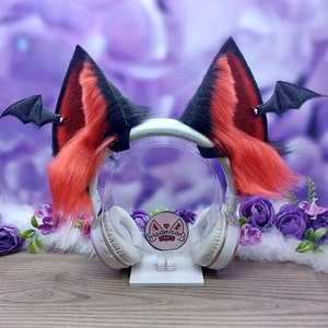 Puede incluir: Orejas de gato de pelo rojo y negro con alas de murciélago unidas a una diadema blanca. La diadema está en un soporte blanco con una pegatina rosa que dice "Meowmeow" con un corazón.