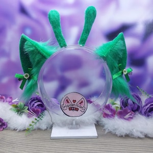 May include: A green faux fur headband featuring two tall, upright antennae and two pointed animal ears. Each ear is decorated with a small green ribbon bow and a central rhinestone. A circular logo with the text Haiamisan is visible.