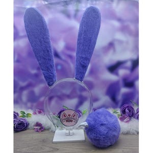 Pode incluir: Um conjunto de orelhas de coelho em pele sintética roxa presas a uma bandolete circular transparente, acompanhado por uma cauda de pompom em pele sintética roxa a combinar. A bandolete apresenta um logótipo circular com o texto Haiamisan.