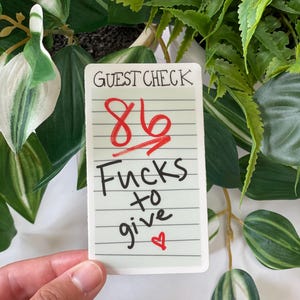 May include: A rectangular sticker with the words "GUEST CHECK" at the top. Below, the number "86" is crossed out in red, with the words "Fucks to give" and a heart symbol. The sticker is held against a background of green plants.
