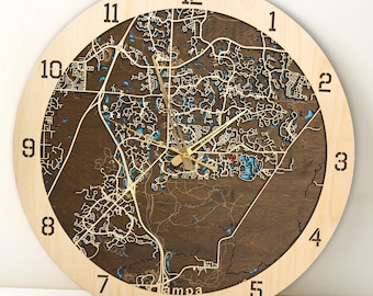 May include: A wooden clock with a brown wood face and a light wood frame. The clock face features a detailed map of Tampa, Florida, with the numbers 1 through 12 around the edge. The clock hands are gold.