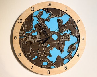 May include: A round wooden clock with a map design. The clock face features a detailed map with blue water areas and brown landmasses. The clock hands are black, and the numbers are in a simple, black font. The outer frame is light-colored wood.