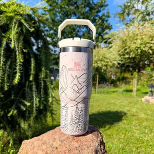 Puede incluir: Una taza de viaje Stanley de acero inoxidable rosa con tapa y asa blancas. La taza presenta una escena de montaña en blanco y negro con la palabra "STANLEY" impresa en el lateral.