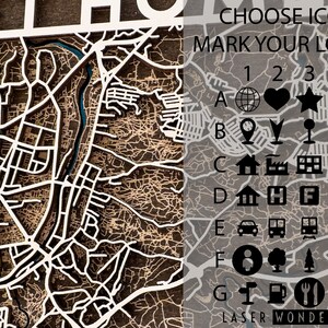 May include: A laser-cut wooden map with a white outline of a city. The map has a red house icon in the center. On the right side of the map, there are icons to mark locations, labeled A through G and 1 through 6. The text "CHOOSE ICONS TO MARK YOUR LOCATIONS" is at the top of the icon section. The text "LASER WONDER THINGS" is at the bottom of the icon section.